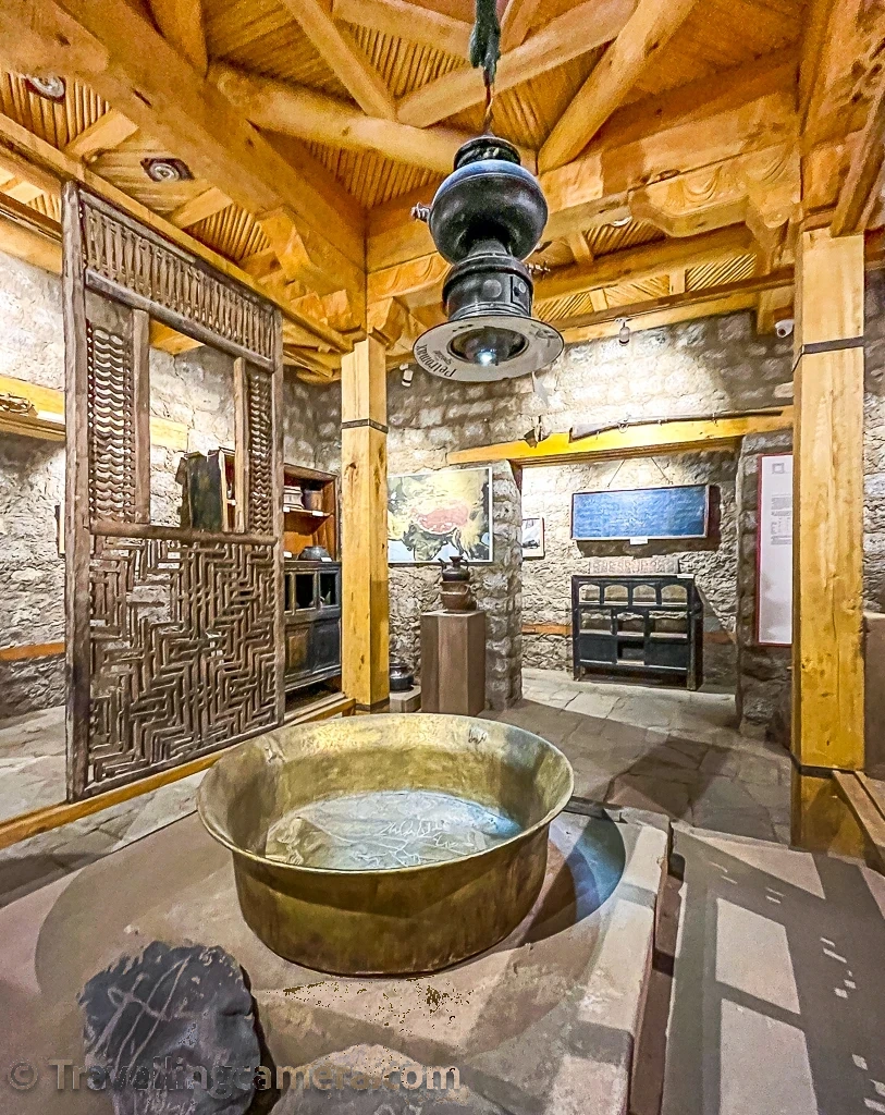 Ground floor gallery with a large brass basin on a stone plinth beneath a hanging metal lamp, a carved wooden lattice screen and stone walls
